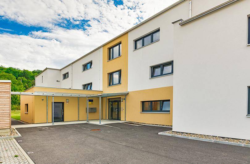 Eingangsbereich eines modernen Wohngebäudes mit überdachtem Zugang und Parkplätzen davor.