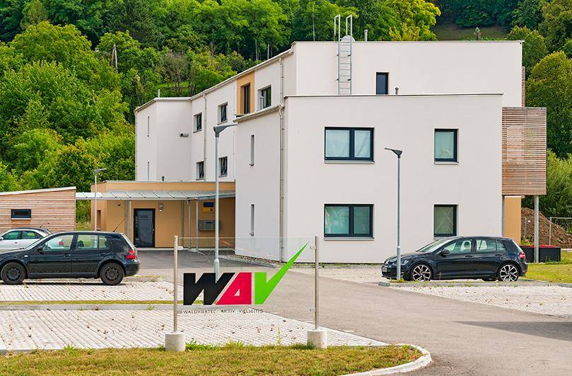 Modernes Wohngebäude mit Parkplätzen und grüner Hügellandschaft im Hintergrund.