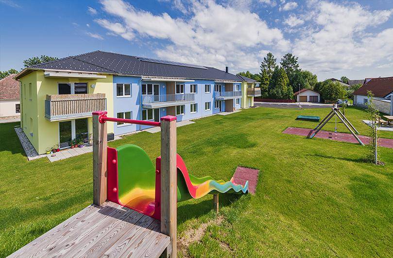 Mehrfamilienhaus mit Spielplatz und Rutsche im grünen Gartenbereich, umgeben von ländlicher Umgebung.