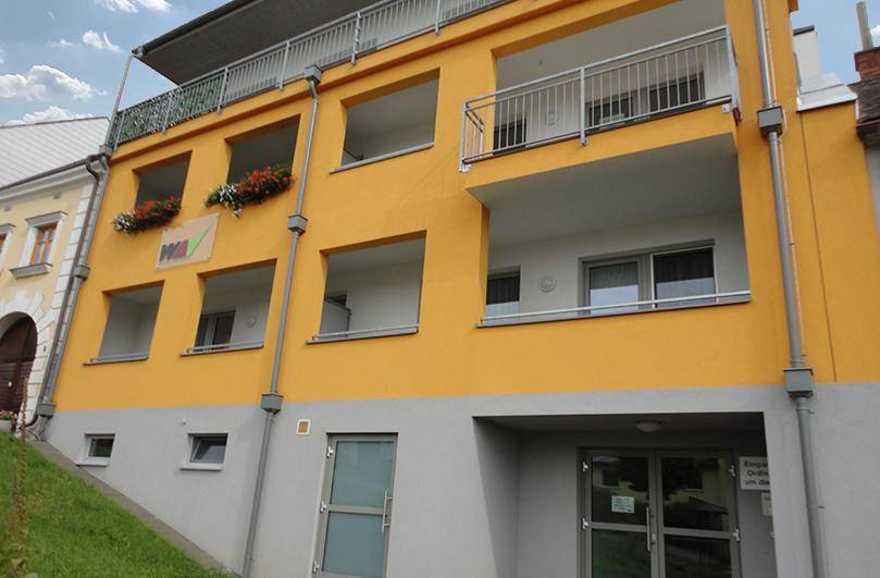 Seitenansicht des Gebäudes mit gelber Fassade, Balkonen und Zugang zum Erdgeschossbereich.