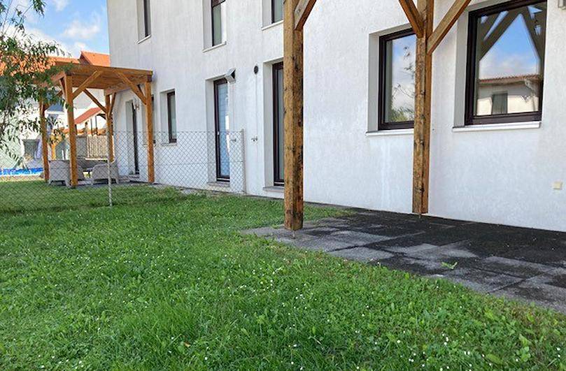 Seitlicher Blick auf das Reihenhaus mit gepflasterter Terrasse unter einer Holzpergola und Rasenfläche.