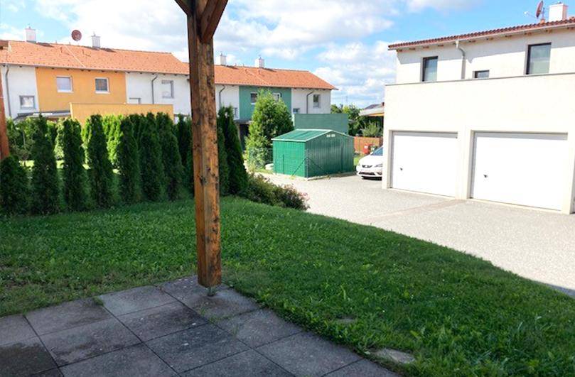 Hinterhofbereich mit Rasen, grünem Gartenschuppen und Doppelgarage, umgeben von Hecken.