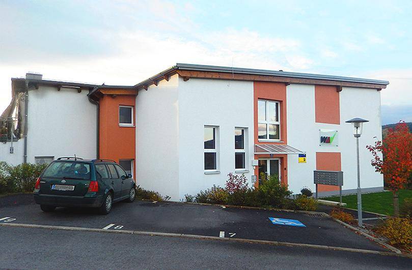 Gebäudefassade mit Parkplätzen und einem Auto vor dem barrierefreien Eingangsbereich.