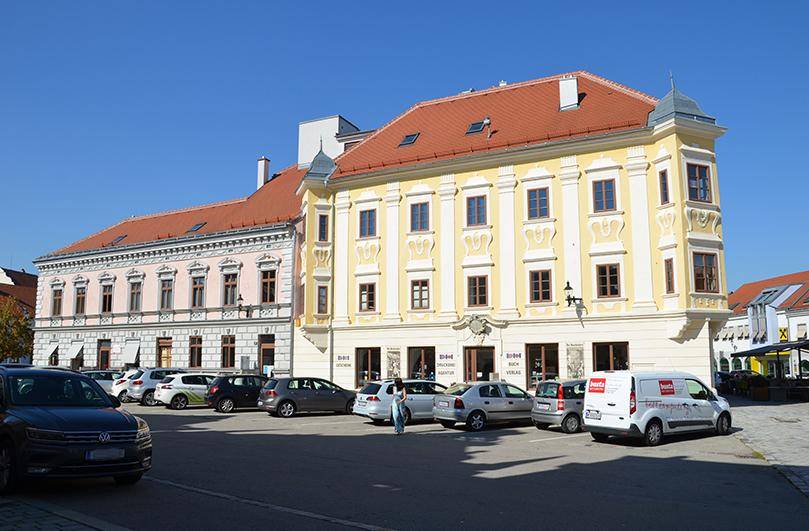 Ansicht eines historischen Gebäudes mit gelber Fassade und geparkten Autos auf dem belebten Stadtplatz.