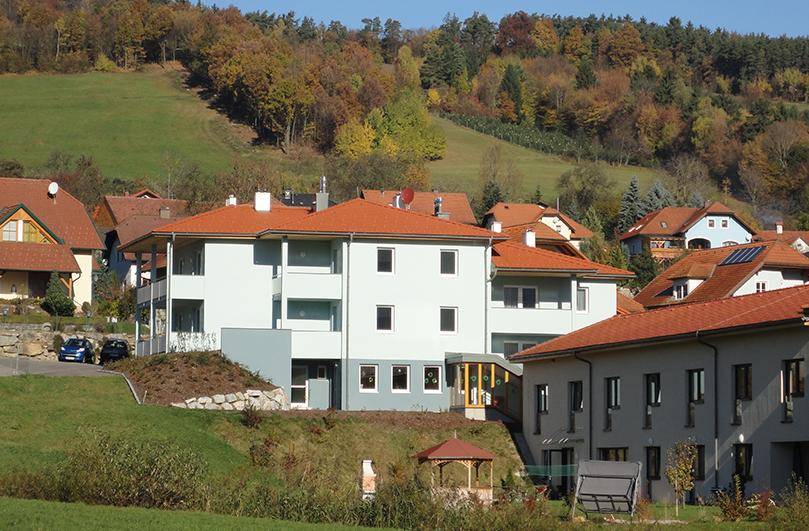Wohnanlage mit roten Ziegeldächern in einer grünen, hügeligen Landschaft mit Herbstbäumen.