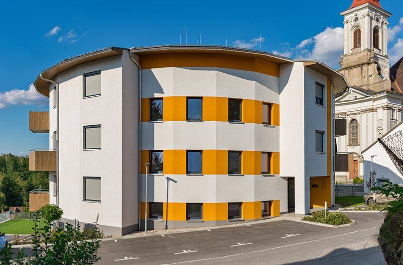 Modernes Mehrfamilienhaus mit runder Architektur, weißen und orangefarbenen Fassadenstreifen sowie Balkonen.