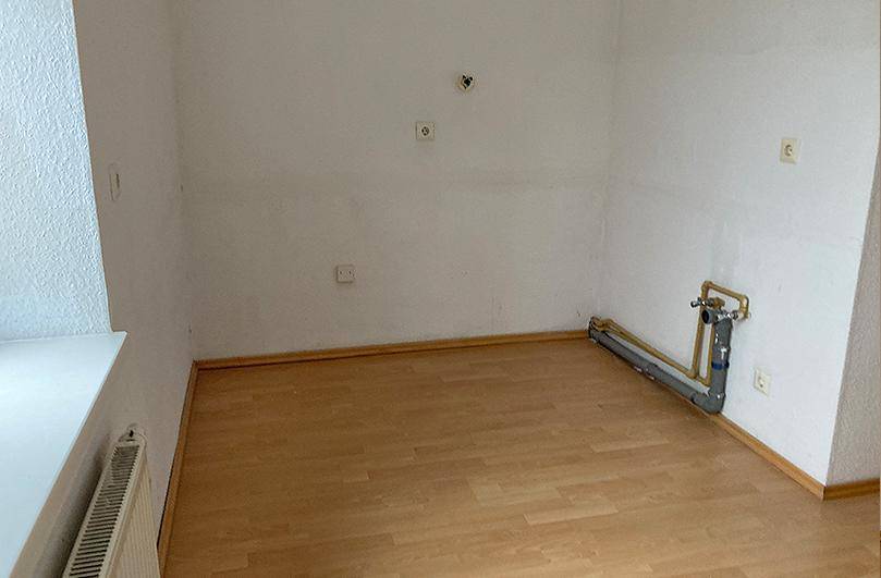 Leerer Küchenraum mit sichtbaren Wasseranschlüssen und Laminatboden, bereit für eine Einbauküche.