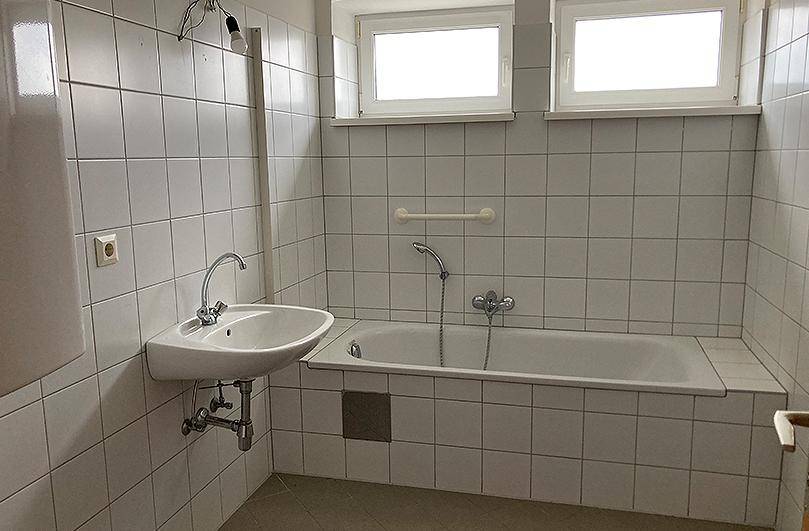 Funktionales Badezimmer mit weißen Fliesen, Badewanne, Waschbecken und zwei Fenstern für Tageslicht.