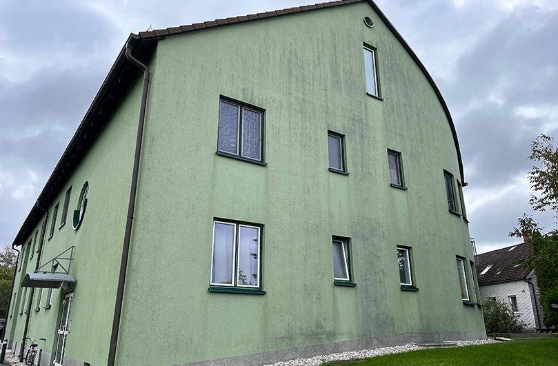 Seitenansicht eines Gebäudes mit grüner Fassade und Satteldach unter bewölktem Himmel.