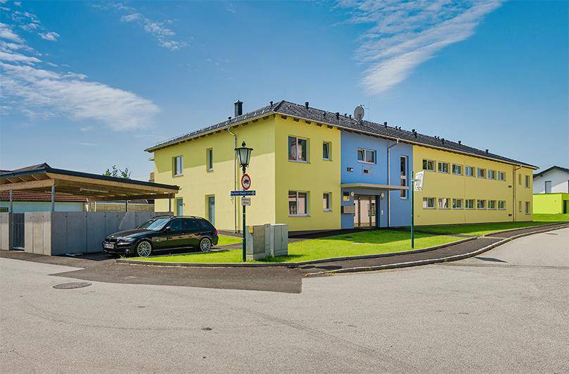 Modernes Mehrfamilienhaus mit gelber und blauer Fassade, Carport und Parkplätzen an einer ruhigen Straße.