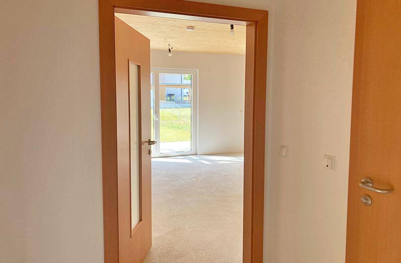 Blick durch eine Tür in einen hellen, leeren Raum mit Holzbalkendecke und Zugang zum Garten.