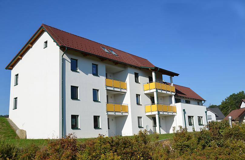 Modernes Wohngebäude mit weißer Fassade, gelben Balkonen und rotem Ziegeldach, eingebettet in eine grüne Umgebung.