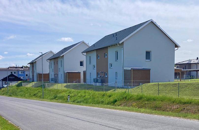 Reihe von modernen Neubauten entlang einer Straße mit grünen Hügeln im Hintergrund.
