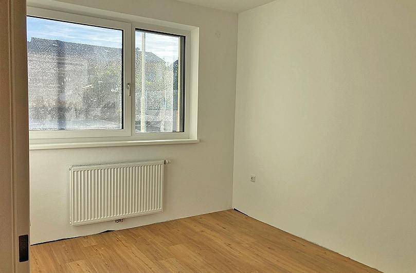Leeres Zimmer mit großem Fenster, Heizkörper und hellem Holzfußboden in neuwertigem Zustand.