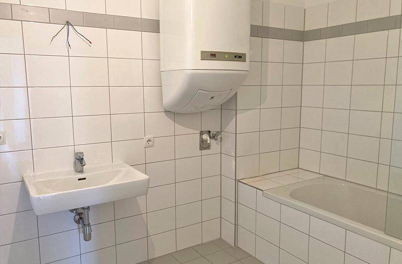 Badezimmer mit weißen Fliesen, Badewanne, Waschbecken und einem Warmwasserboiler an der Wand.