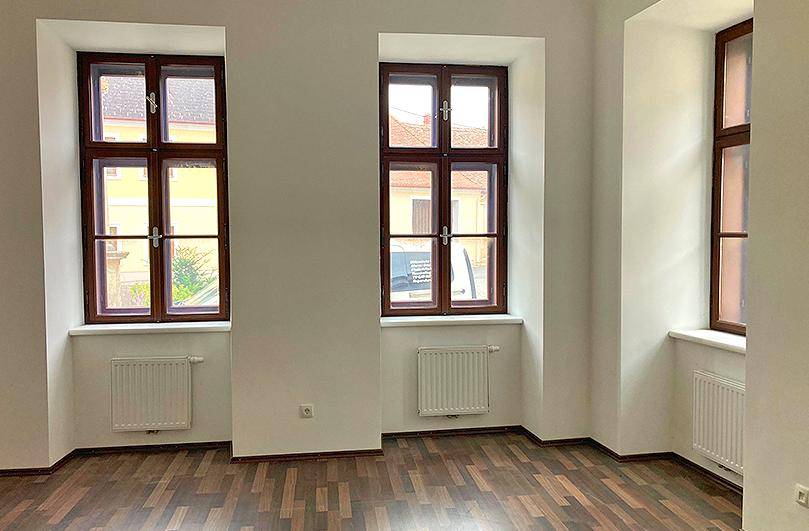 Heller Raum mit zwei großen Fenstern, dunklem Laminatboden und weißen Wänden.