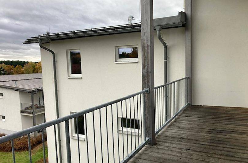Balkon mit Holzbelag und Metallgeländer, Blick auf die Gebäudeseite und Herbstbäume.