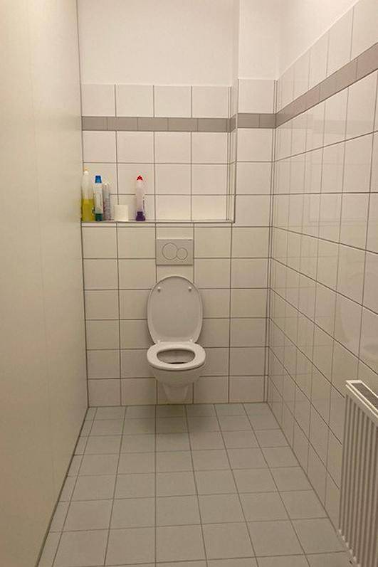 Einfaches WC mit weißen Fliesen, wandmontierter Toilette und einem kleinen Regal.