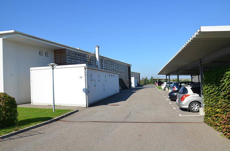 Außenansicht der Wohnanlage mit überdachten Parkplätzen und Zufahrt unter blauem Himmel.