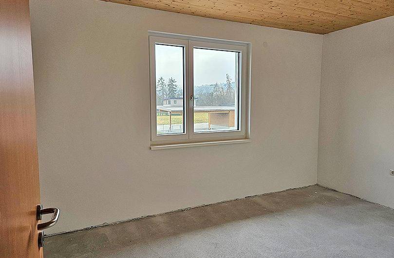 Schlafzimmer im Rohbau mit Holzdecke, Fenster und Blick auf die Umgebung.