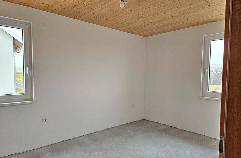 Schlafzimmer im Rohbau mit Holzdecke, großen Fenstern und Blick ins Grüne.