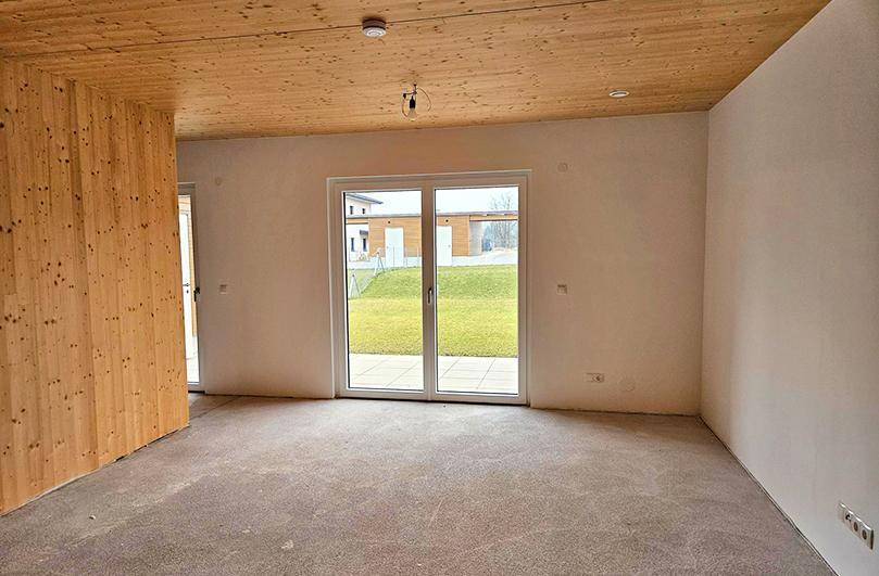 Geräumiges Wohnzimmer im Rohbau mit Holzwand, Holzdecke und Zugang zum Garten.