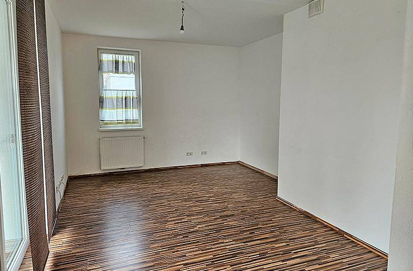 Leeres Wohnzimmer mit Laminatboden, weiß gestrichenen Wänden und einem Fenster mit Jalousien.