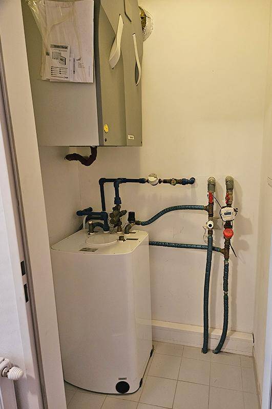 Technikraum mit Warmwasserboiler und sichtbaren Rohrleitungen auf gefliestem Boden.