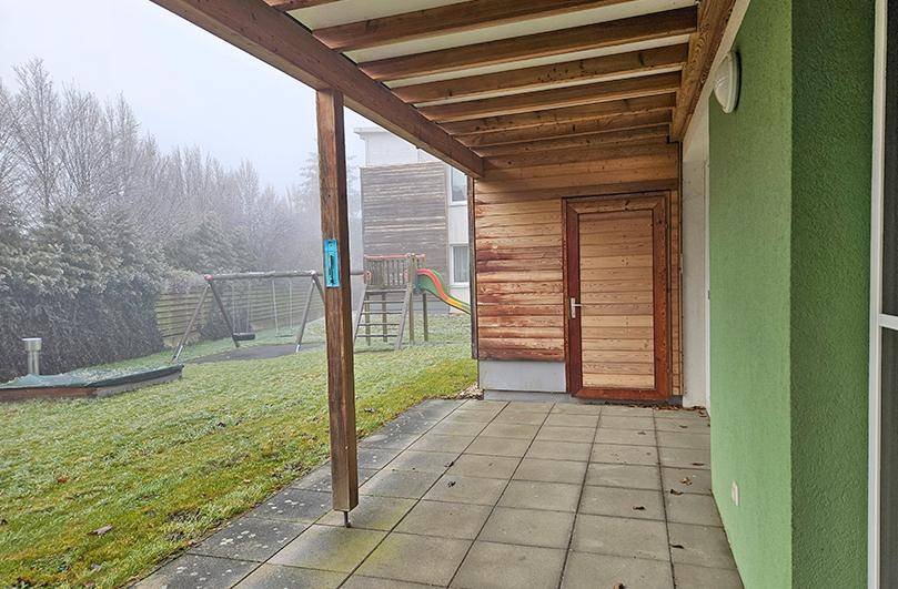 Überdachte Terrasse mit Fliesenboden und Blick auf einen Spielplatz im Garten.