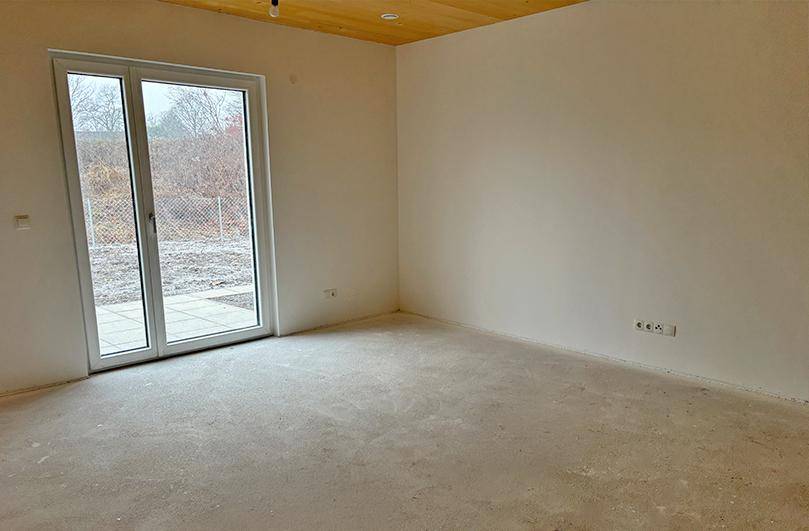 Unfertiger Wohnraum mit Betonboden, hellen Wänden, Holzdecke und großer Terrassentür.