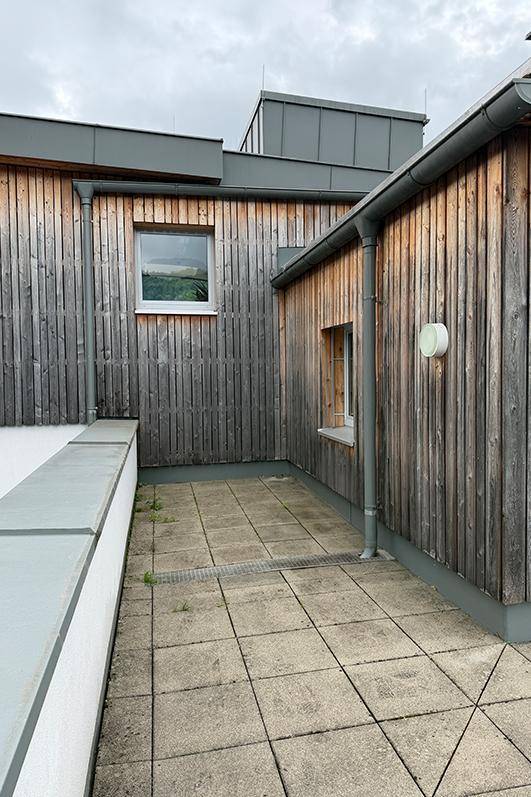 Außenbereich mit Holzfassade und gefliester Terrasse, umgeben von einer schützenden Brüstung.