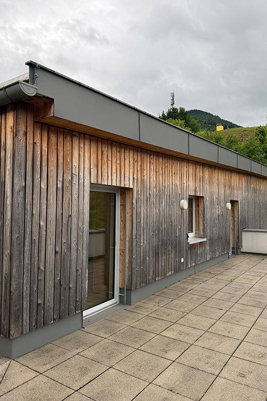 Holzverkleidete Fassade mit großen Fenstern und Zugang zu einer weitläufigen, gefliesten Terrasse.