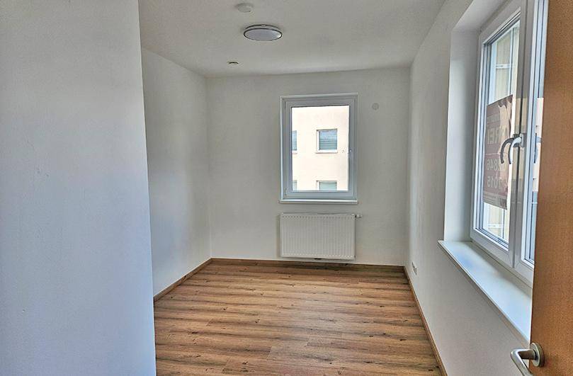 Helles Zimmer mit Laminatboden und einem Fenster, das den Blick auf ein gegenüberliegendes Gebäude freigibt.