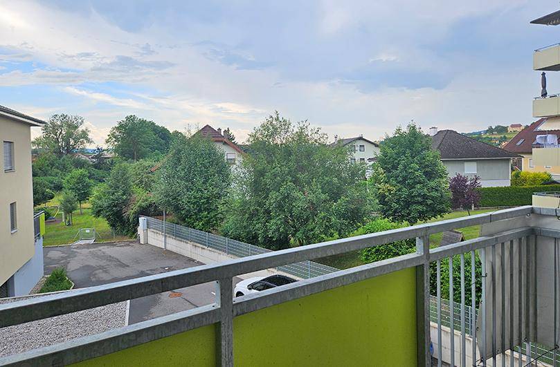 Balkon mit Metallgeländer und Blick auf eine grüne Umgebung mit Bäumen und Häusern.