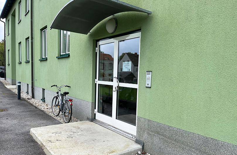 Eingangsbereich des grünen Gebäudes mit moderner Glastür, Gegensprechanlage und Vordach.
