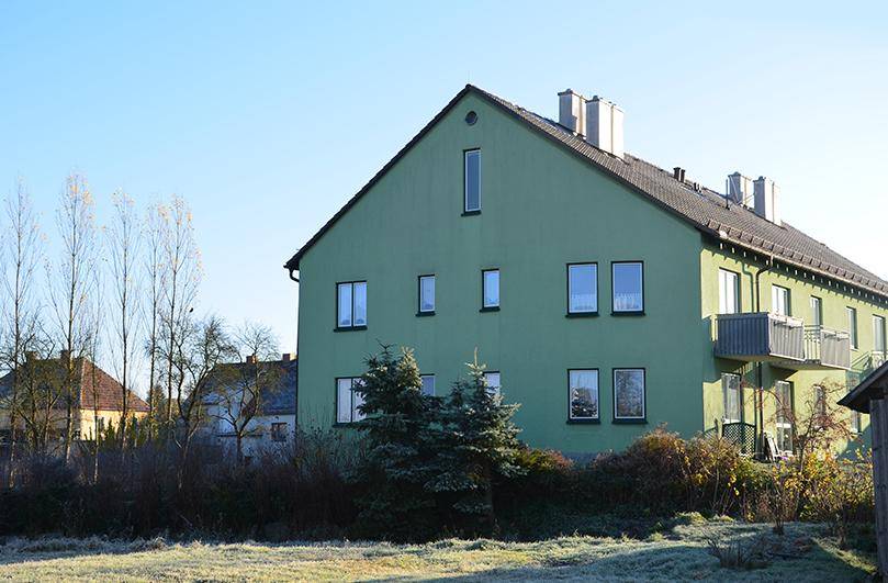 Grünes Wohngebäude mit Satteldach und Balkonen, umgeben von einer ländlichen Landschaft unter blauem Himmel.