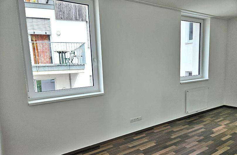 Heller Raum mit Laminatboden und Fenstern, die einen Blick auf einen gegenüberliegenden Balkon bieten.