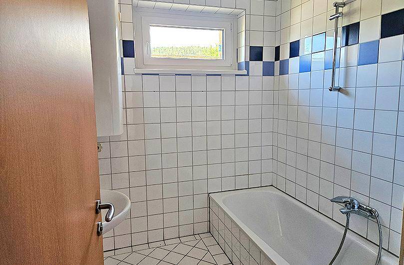 Funktionales Badezimmer mit Badewanne, Dusche und Fenster, gefliest in Weiß und Blau im Stil der 80er Jahre.
