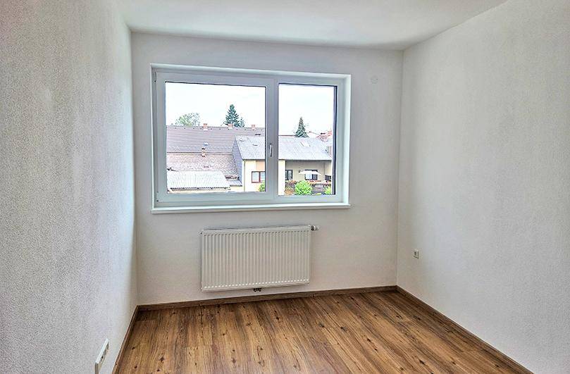 Leeres Zimmer mit Laminatboden, weiß gestrichenen Wänden und einem Fenster mit Blick auf die Dächer.