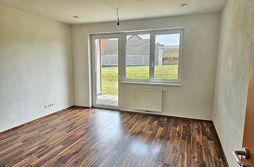 Leerer Raum mit Laminatboden, großem Fenster und Zugang zu einer Terrasse oder Garten.