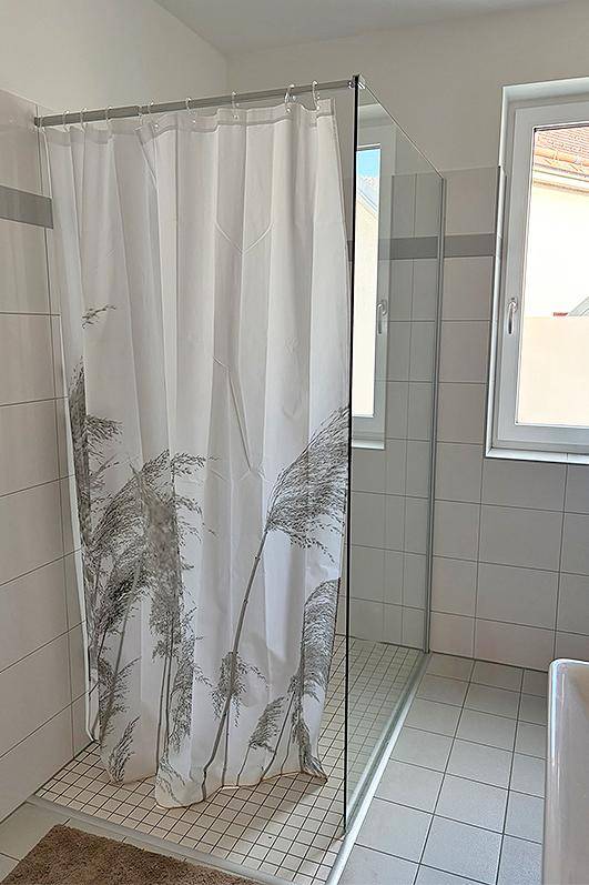 Duschbereich im Badezimmer mit Duschvorhang, Fliesenboden und Fenster für Tageslicht.