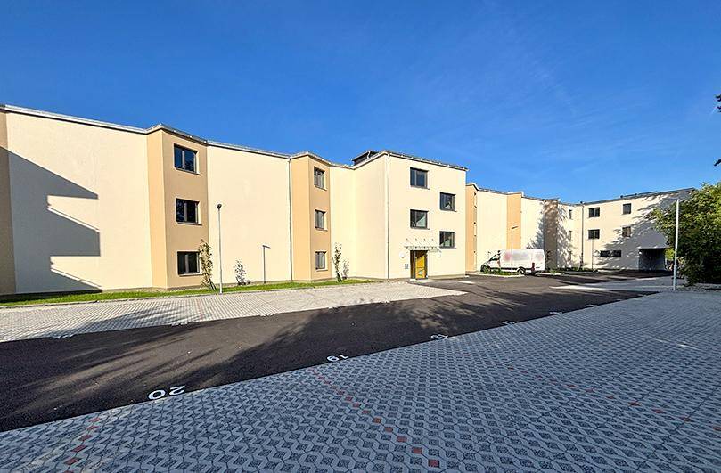 Rückseite des Mehrfamilienhauses mit Außenparkplätzen und gepflastertem Bereich unter blauem Himmel.