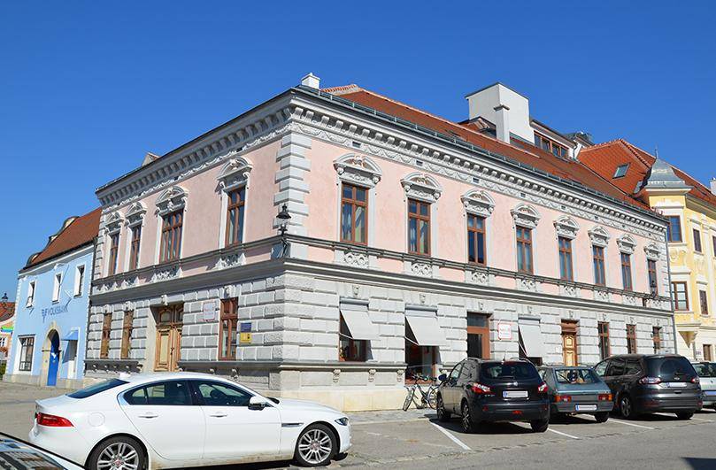 Historisches Eckgebäude mit rosa Fassade, vielen Fenstern und traditionellem Ziegeldach an einem sonnigen Tag.