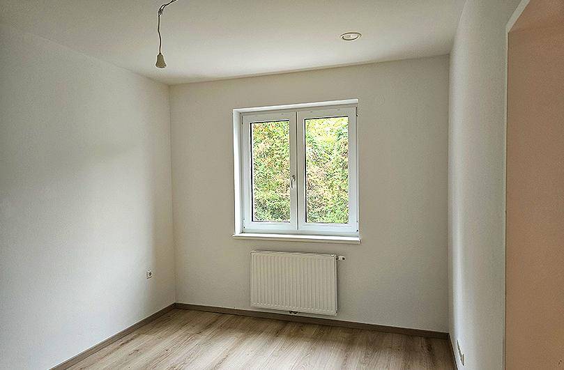 Helles Zimmer mit Fenster und Blick ins Grüne, ausgestattet mit Heizkörper und Laminatboden.