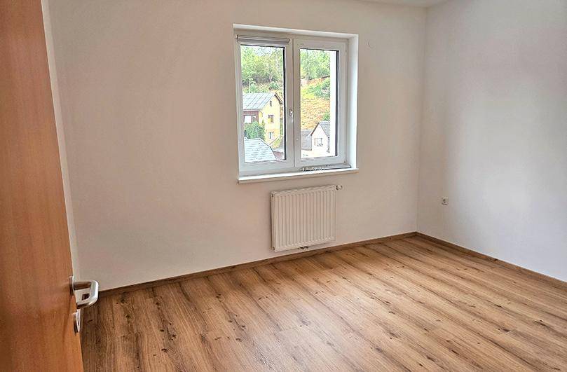 Leeres Schlafzimmer mit Laminatboden, weiß gestrichenen Wänden und einem Fenster mit Blick ins Grüne.