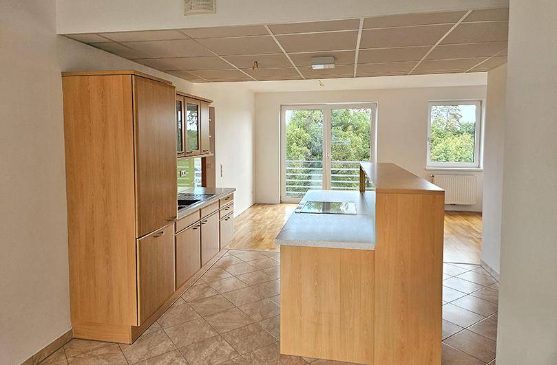 Blick von der Küche in den hellen Wohnbereich mit Holzboden und Zugang zu einem Balkon mit Grünblick.