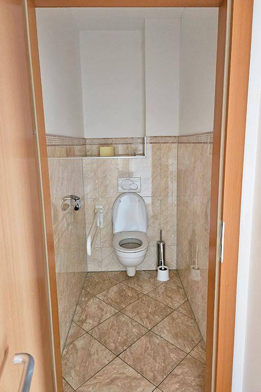 Separates WC mit beigefarbenen Fliesen, wandhängender Toilette und Haltegriffen für zusätzlichen Komfort.