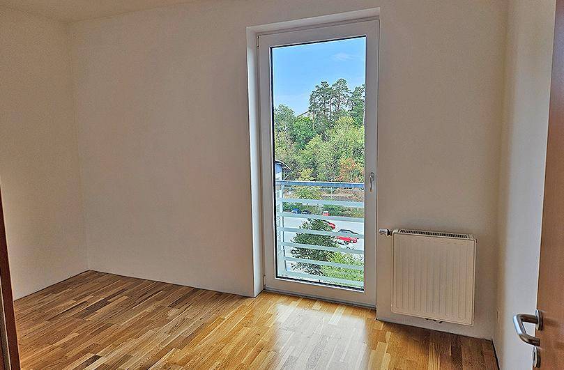 Freundliches Zimmer mit Holzboden und großem Fenster, das einen grünen Ausblick und viel Tageslicht bietet.