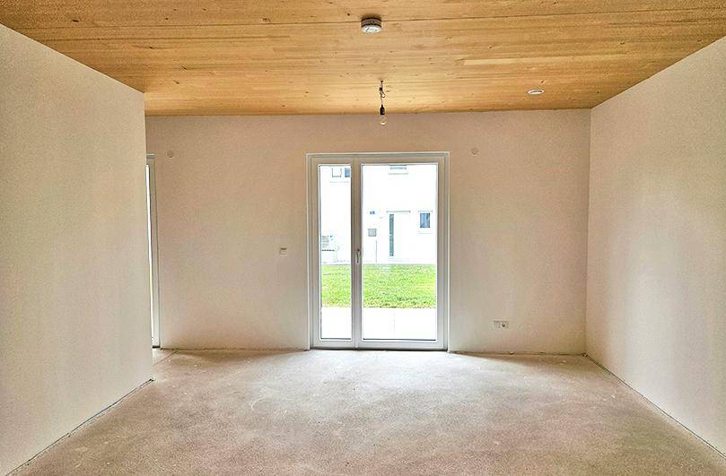 Heller, unmöblierter Wohnraum mit Holzbalkendecke und Zugang zu einer Rasenfläche.