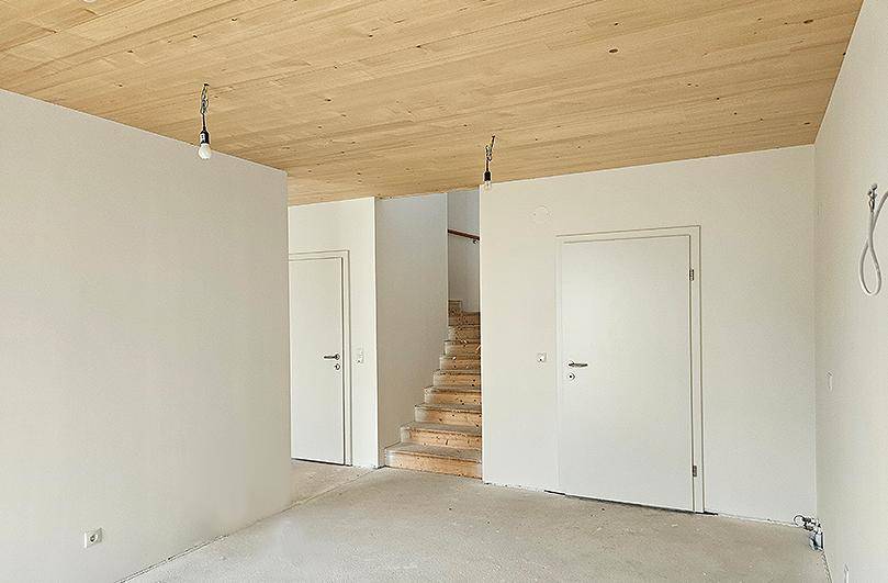 Geräumiger, unmöblierter Raum mit Holzbalkendecke und offener Holztreppe ins Obergeschoss.